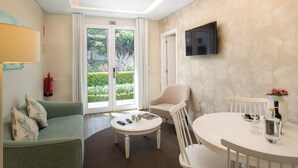 Une chambre d’hôtel avec un canapé vert, une table blanche avec des télécommandes, une chaise blanche, un téléviseur fixé au mur et une porte donnant sur un jardin.