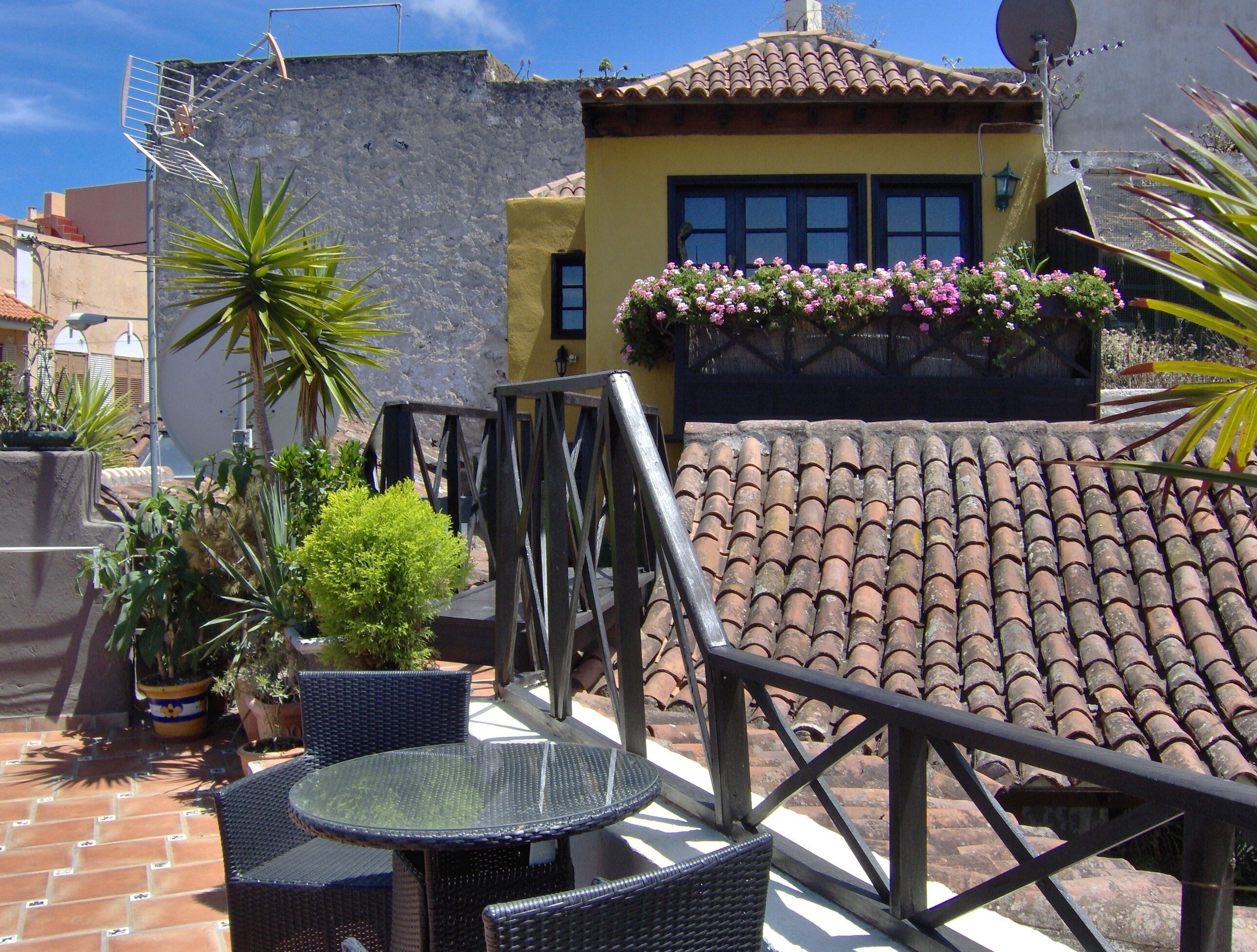 A patio with a round glass table and wicker chairs, surrounded by potted plants and a small garden.