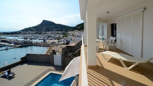 Un balcón con piscina, muebles blancos y vistas a un puerto deportivo y un pueblo en la colina.