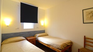 A room with two single beds, a small wooden nightstand, a window with a black blind, and wall-mounted lights.