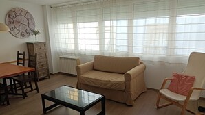 A living room with a wooden table, a beige sofa, a glass coffee table, a wooden chair, and a clock on the wall.