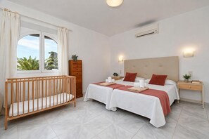A hotel room with a crib, a double bed with red pillows, a nightstand, and a window with curtains.