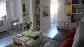 Una sala de estar con un sofá gris, una mesa de centro con libros y un florero, una estantería blanca y una cocina al fondo.