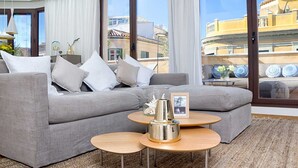 A modern living room with a grey sectional sofa, two round wooden tables, and a view of buildings through large windows. - Kim - Holiday Penthouse Malaga City (Málaga)