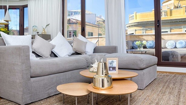 A modern living room with a grey sectional sofa, two round wooden tables, and a view of buildings through large windows.