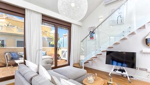 A modern living room with a grey sofa, a flat-screen TV, and a staircase with glass railings. - Kim - Holiday Penthouse Malaga City (Málaga)