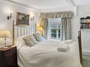 A neatly arranged bedroom with a bed, bedside lamps, a painting, a window with curtains, and a bookshelf.