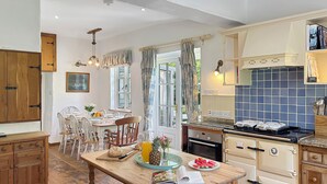 A kitchen with wooden cabinets, a dining table with chairs, a stove, and a window with curtains. - Bratton Mill Farmhouse, Bratton Fleming (Bratton Fleming)