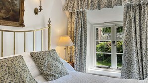 A bedroom with a bed, a nightstand, a lamp, a window with curtains, and a painting on the wall. - Bratton Mill Farmhouse, Bratton Fleming (Bratton Fleming)