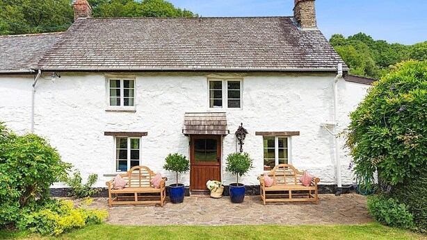 A white house with a tiled roof, a wooden door, and two windows on the front. There are two wooden benches with cushions and potted plants in front. - Bratton Mill Farmhouse, Bratton Fleming (Bratton Fleming)