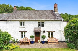 A white house with a tiled roof, a wooden door, and two windows on the front. There are two wooden benches with cushions and potted plants in front. - Bratton Mill Farmhouse, Bratton Fleming (Bratton Fleming)
