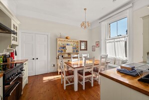 A kitchen with white cabinetry, a wooden dining table, and chairs. There is a built-in shelf with dishes and a hanging light fixture.