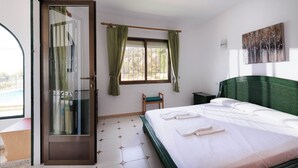 Une chambre d’hôtel avec un lit, une chaise, une fenêtre avec des rideaux, un tableau au mur et une porte coulissante donnant sur un espace extérieur.