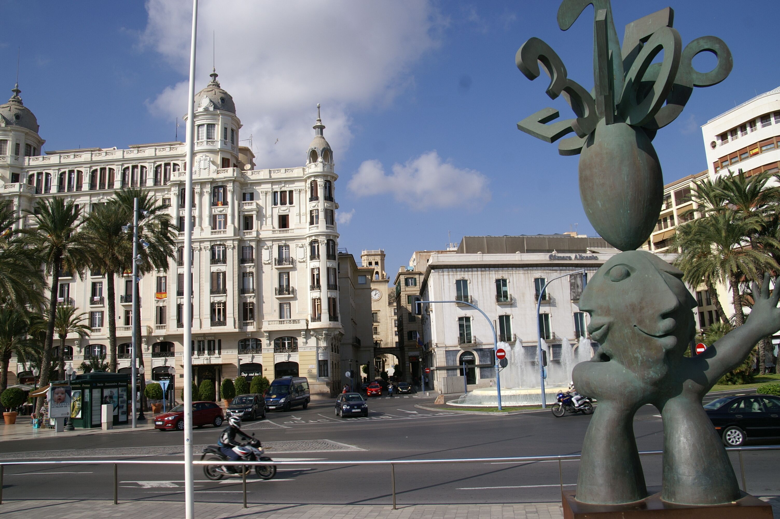 Una calle con un gran edificio de arquitectura clásica, palmeras y una escultura.