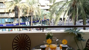 Un balcone con un tavolo apparecchiato per la colazione, con vista su una strada alberata da palme e palazzi.