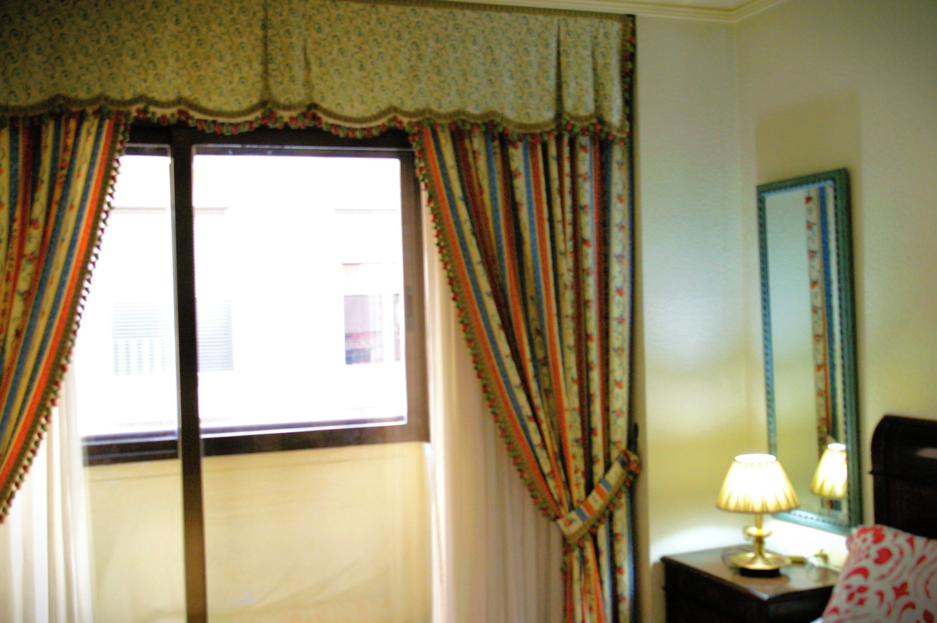 A room with a large window, curtains, a lamp, and a mirror on the wall.