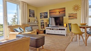 Un salón moderno con sofá, reposapiés y un televisor de pantalla plana sobre un soporte blanco. Hay cuadros en las paredes, una mesa de comedor con sillas y un amplio ventanal con cortinas.