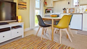 Una cocina moderna con armarios blancos, una mesa de comedor con sillas, un televisor sobre un mueble bajo y una ventana con cortinas.