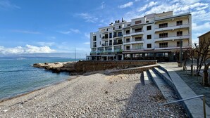 Un edificio costero con restaurante y pizzería en la planta baja, un muro de piedra en primer plano y una playa de guijarros.