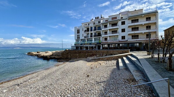 A coastal building with a restaurant and pizzeria on the ground floor, a stone wall in front, and a pebble beach.