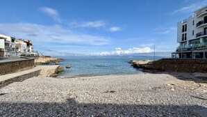 Una playa de guijarros con costa rocosa, cielo azul despejado y edificios a ambos lados.
