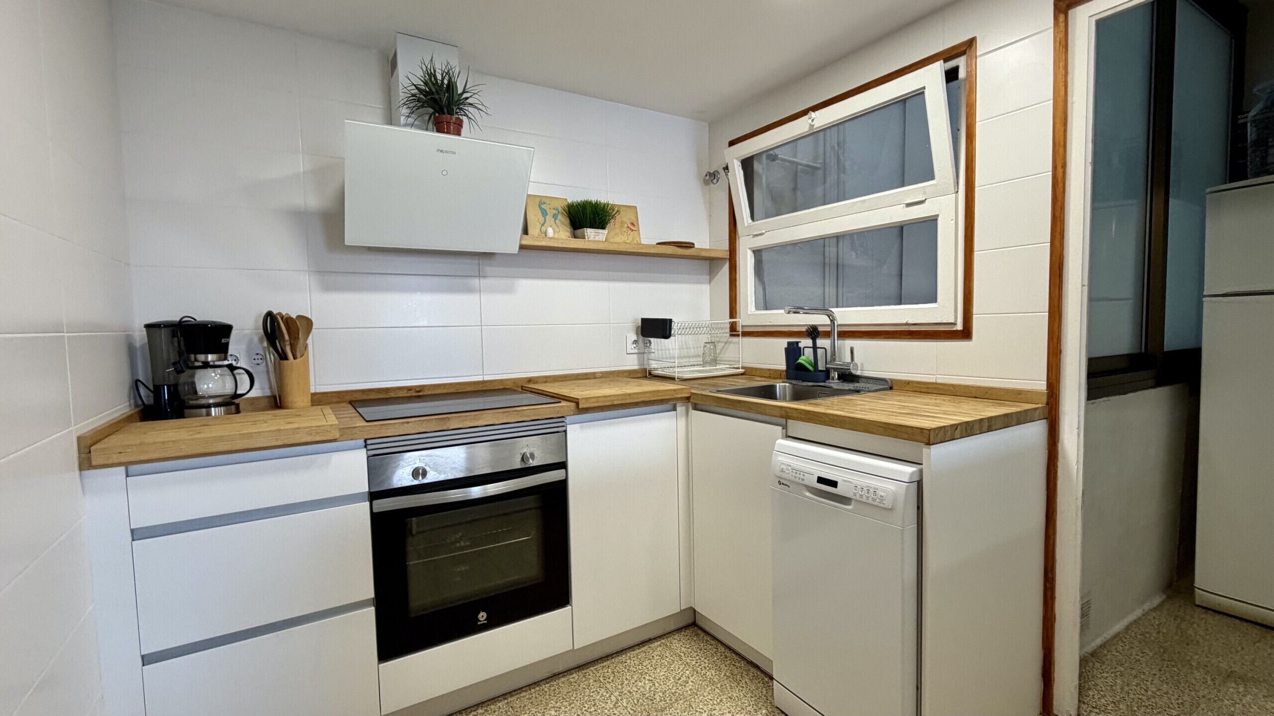 Een functionele keuken met witte kastjes, een ingebouwde oven, een vaatwasser, een koffiezetapparaat en een raam met uitzicht.