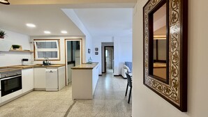 Una cocina moderna con armarios blancos, horno empotrado y lavavajillas. Hay una pequeña isla con un fregadero y una ventana encima. La cocina da a un pasillo con una puerta al final.