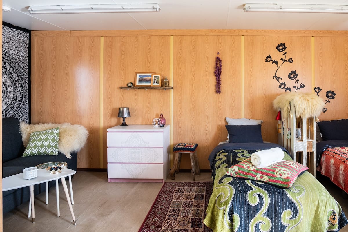 A cozy room with two beds, a small table, a dresser, and a wall-mounted shelf with decorative items.