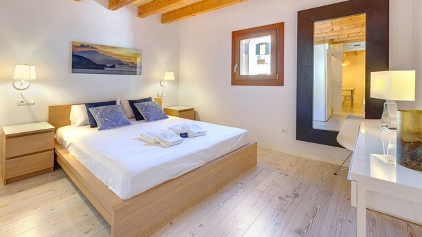 A bedroom with a wooden bed, two bedside tables, a nightstand, a mirror, and a painting on the wall. - URBAN SUITES 2 PALMA STANDARD by PriorityVillas (Palma de Mallorca)