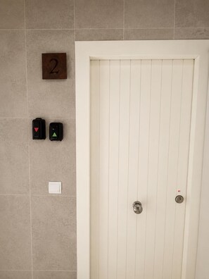 Une porte blanche avec un trou de serrure et une serrure, un numĂ©ro « 2 » fixĂ© au mur, et deux boutons dâurgence avec des symboles rouges et verts.