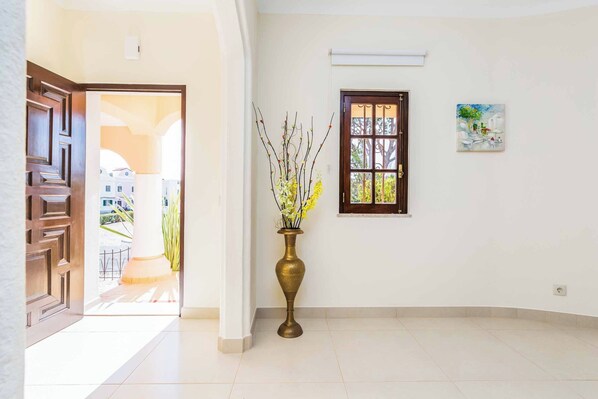 A hallway with a large wooden door, a tall brass vase with yellow flowers, a window with a view of the outside, and a painting on the wall.