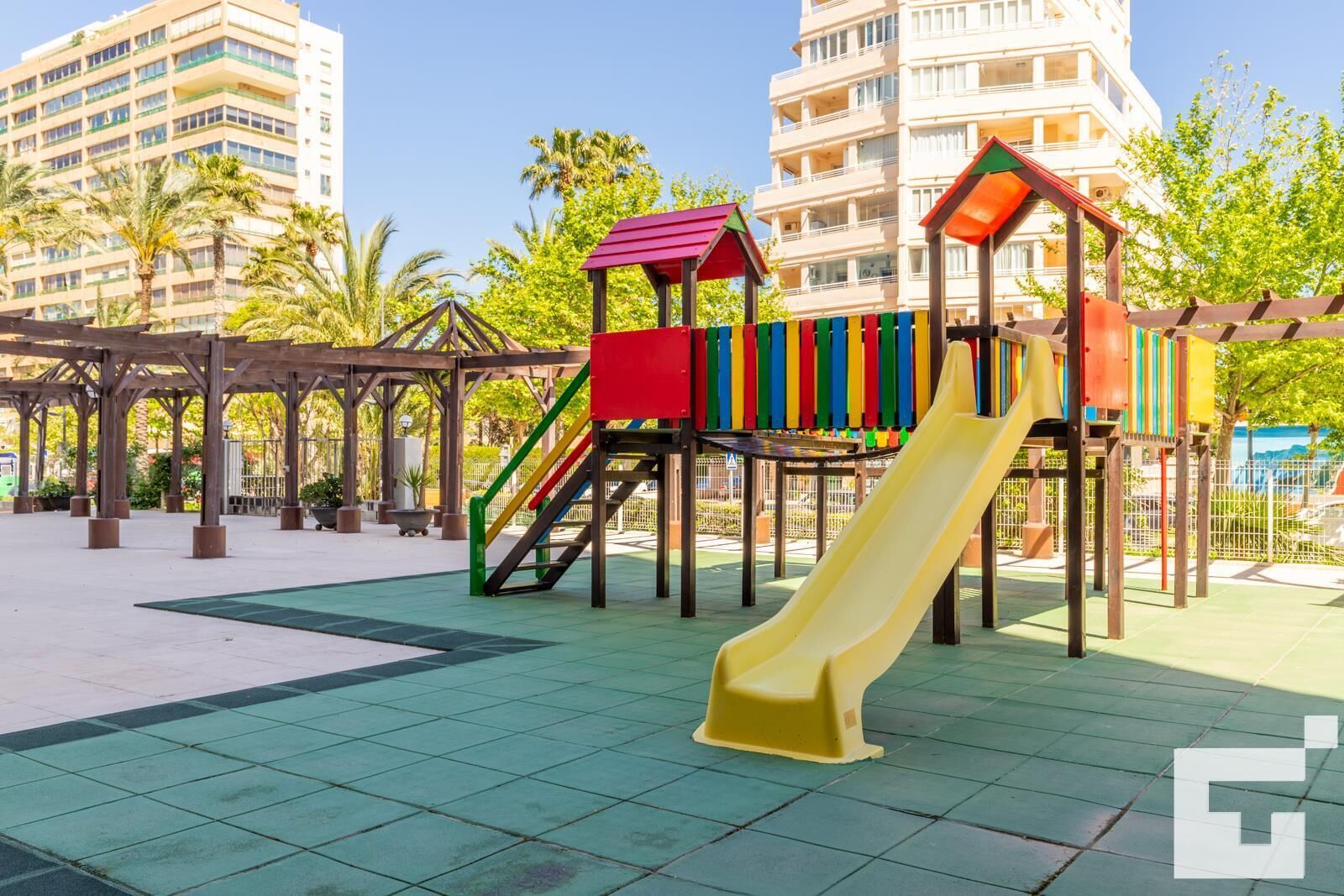 A playground with a slide and climbing structures, surrounded by a wooden pergola and palm trees.