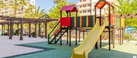 A playground with a slide and climbing structures, surrounded by a wooden pergola and palm trees.