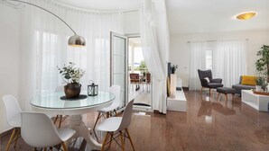 Un salon moderne avec une table à manger en verre, des chaises blanches, un canapé gris et un fauteuil noir. On aperçoit une plante en pot sur le canapé et une lampe sur pied dans un coin.