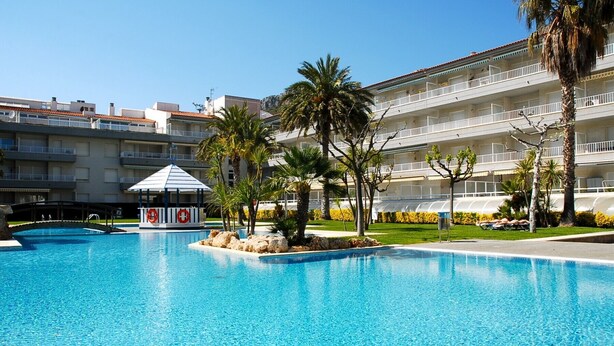 A swimming pool with a lifeguard station, palm trees, and a multi-story hotel building in the background.