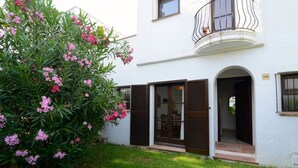 A white building with a balcony, a door with a window, and a small garden with pink flowers. - PIVERD DEL GOLF 56 (Pals)