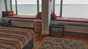 Ett sovrum med stora fönster, en säng, en bänk och utsikt över havet.