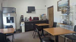 A small kitchenette with a refrigerator, microwave, coffee maker, and a sink. There are two wooden tables and four chairs. A painting of a mountain landscape hangs on the wall. - Western Inn (Green River)