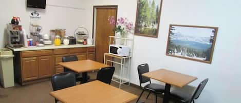A room with a kitchenette, a microwave, a coffee maker, and a refrigerator. There are two tables and four chairs.
