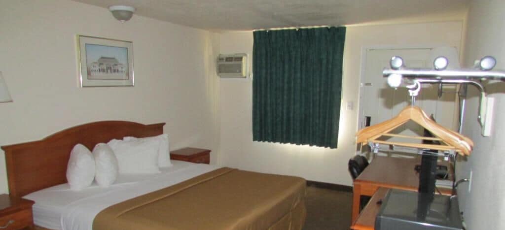 Habitación de hotel con una cama, una mesita de noche de madera, un espejo con luces, una ventana con cortinas y un cuadro en la pared.