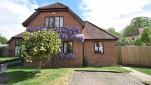 A two-story brick house with a sloped roof, a visible garage door, and a tree with purple flowers in the front yard. - Garden Flat -  comfortable cottage for two close to Chichester and Goodwood (Tangmere)