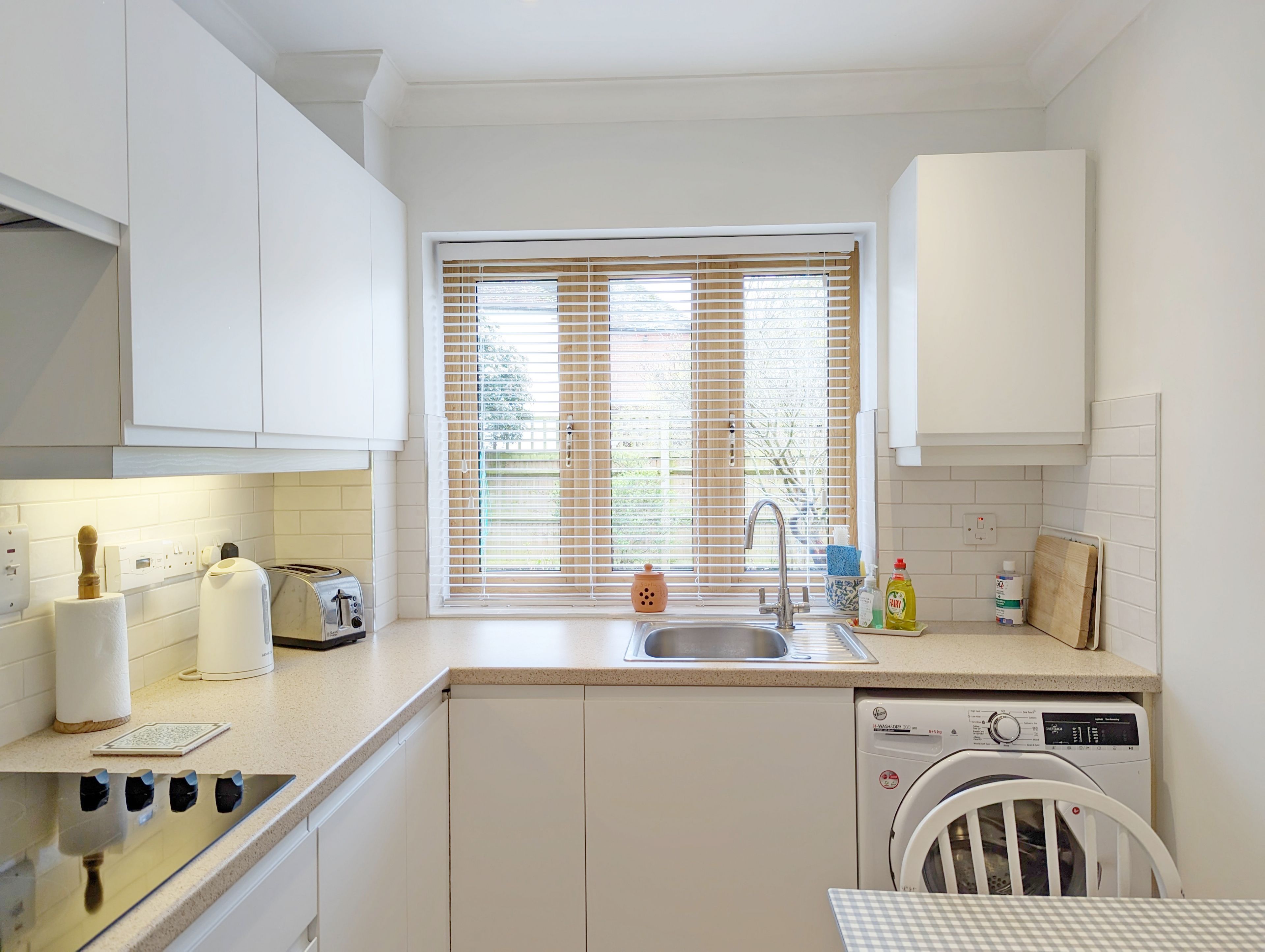 A modern kitchen with white cabinets, a built-in dishwasher, a sink, and a window with blinds.