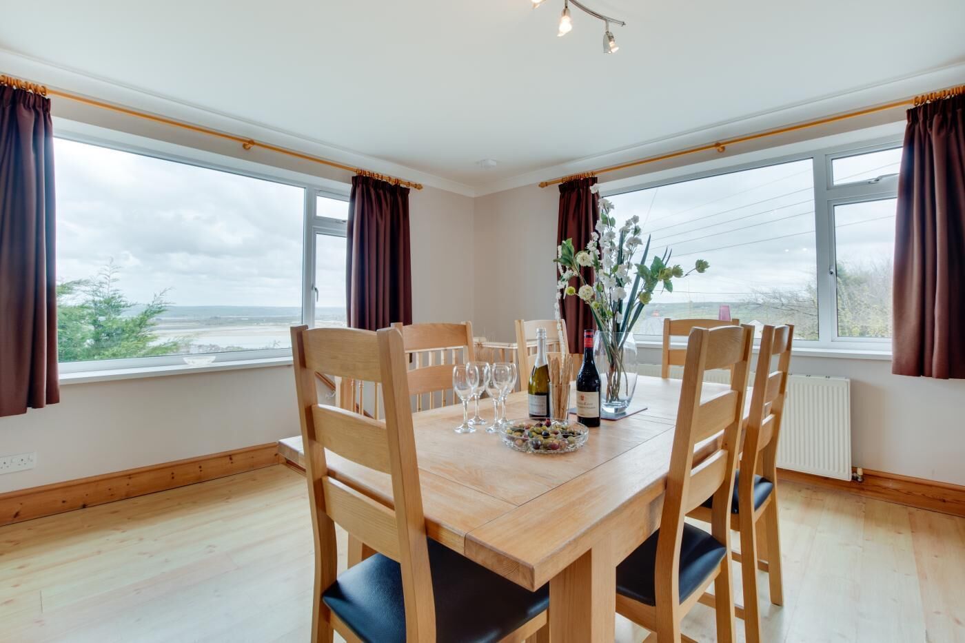 A dining room with a wooden table and chairs, a vase with flowers, and a view of the sea through large windows.
