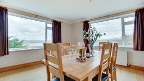 A dining room with a wooden table and chairs, a vase with flowers, and a view of the sea through large windows. - 4 Bed in Appledore (oc-tidal) (Appledore)