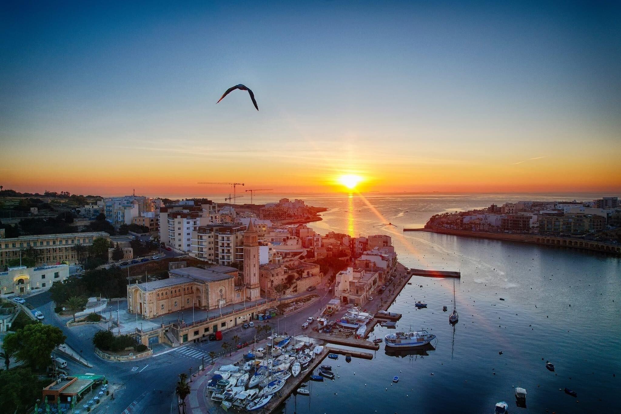 Une vue d’une ville côtière au coucher du soleil, avec un port de plaisance, des bâtiments et des bateaux.