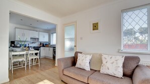 A living room with a beige sofa, a dining table with chairs, and a kitchen in the background.
