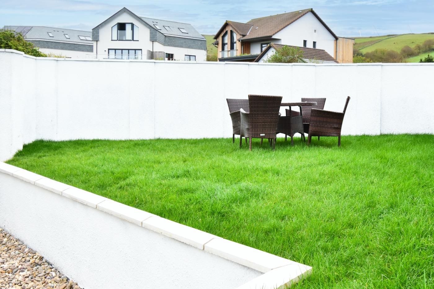 A well-maintained grassy backyard with a white wall, a concrete pathway, and a dining table with chairs.