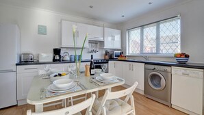 A modern kitchen with white cabinets, a dining table set for four, a washing machine, and a window with blinds.