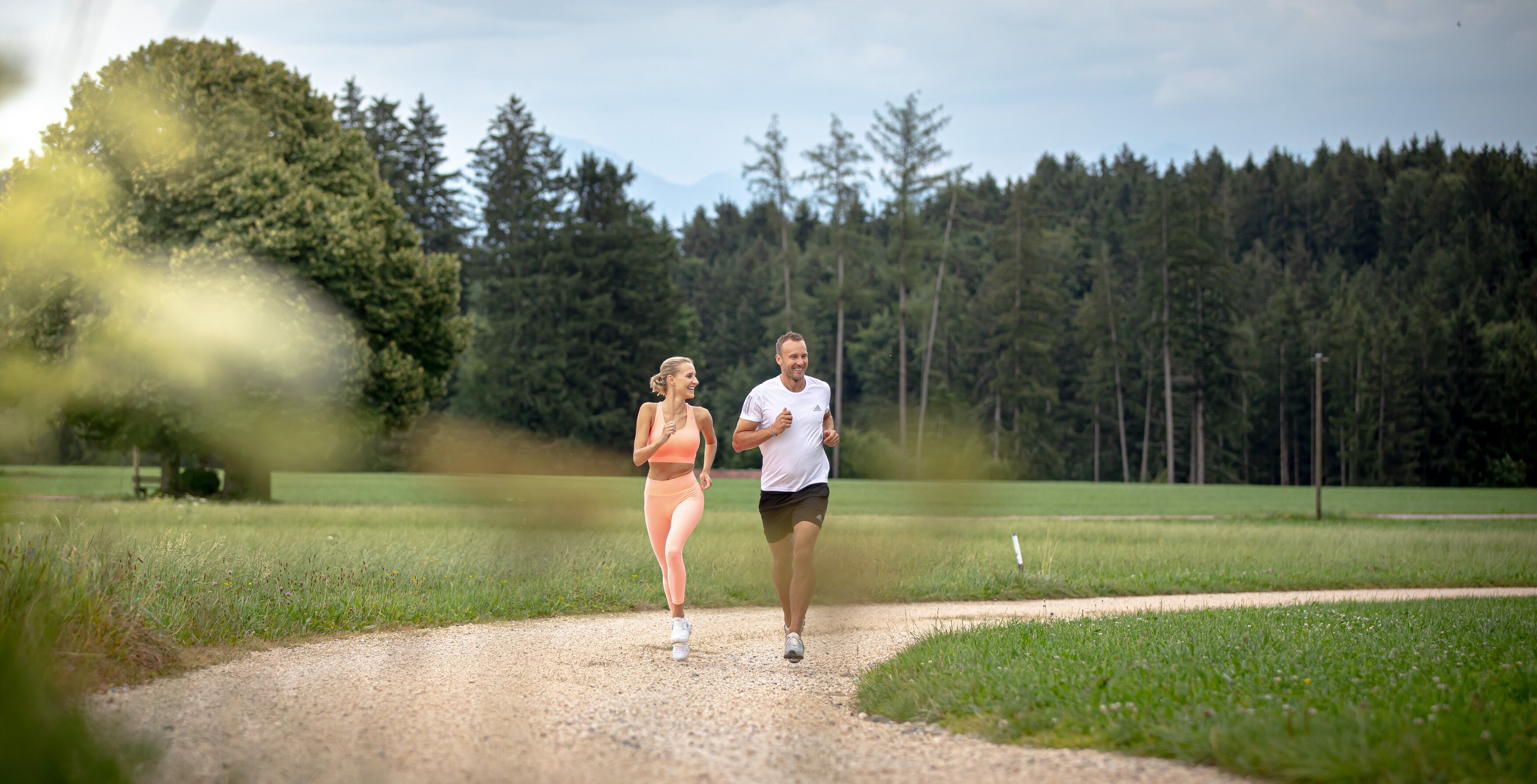Zwei Personen joggen auf einem Kiesweg, umgeben von Grünflächen und Bäumen.