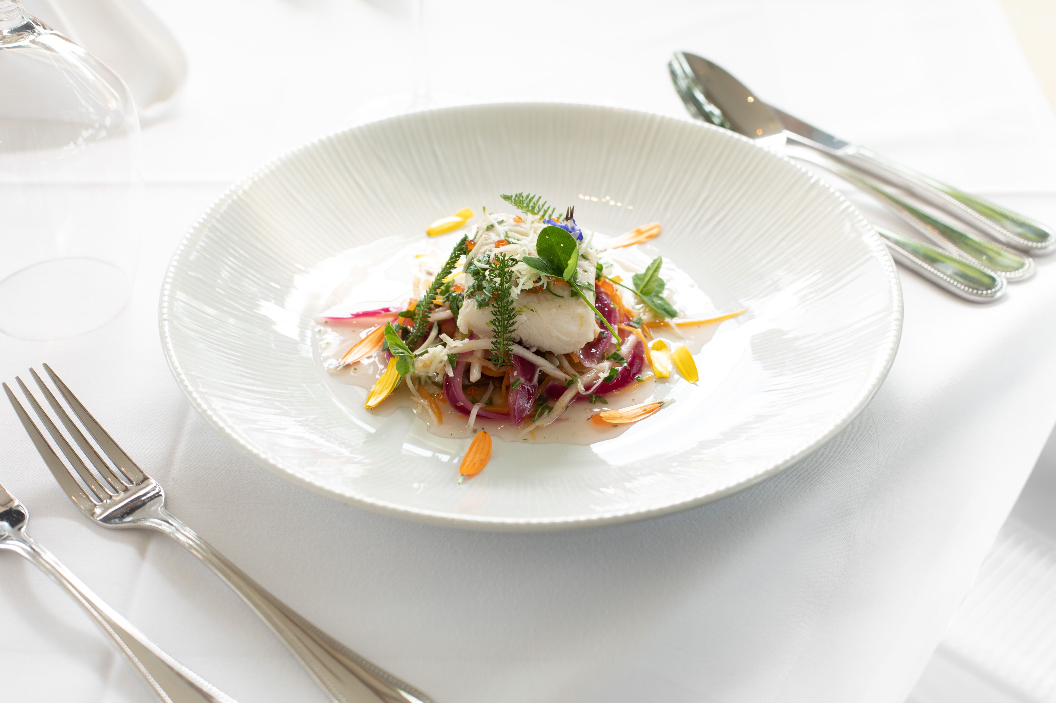 A white plate with a fish dish, garnished with herbs and vegetables, on a white tablecloth.